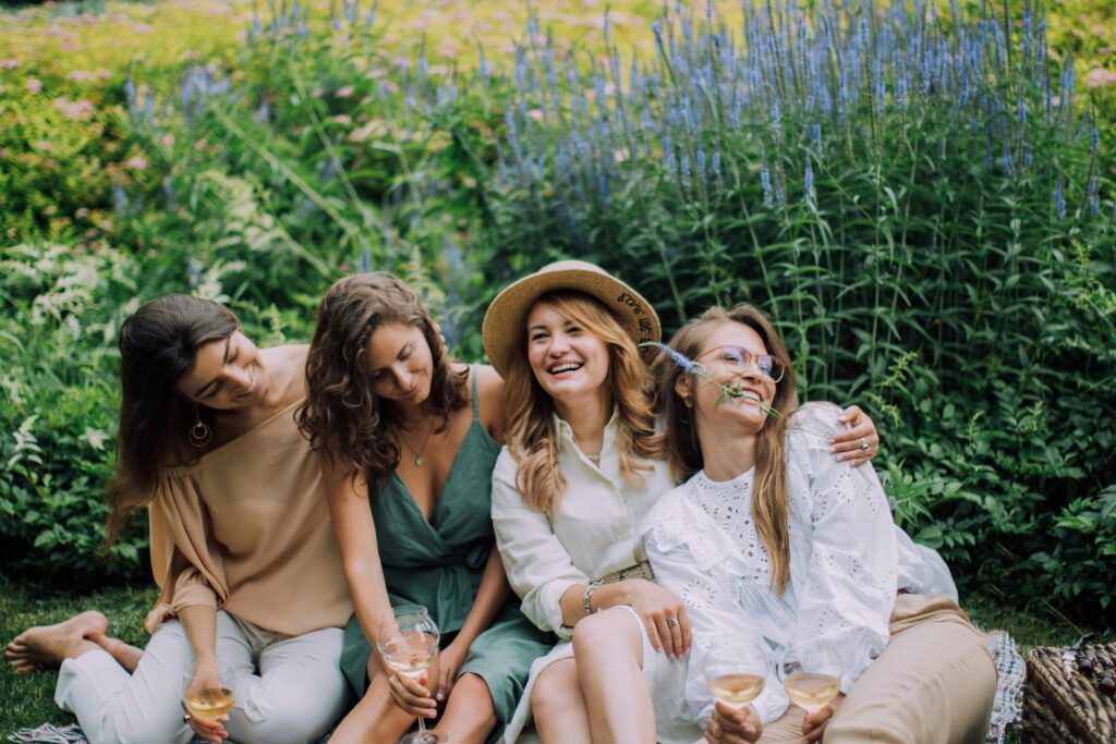 Four women enjoying a cheerful picnic with wine in a vibrant garden setting.