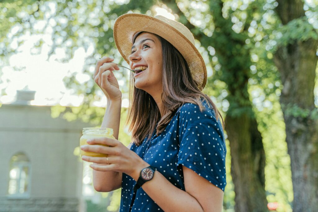 Smiling woman wearing a hat enjoys a dessert outdoors under sunlight.